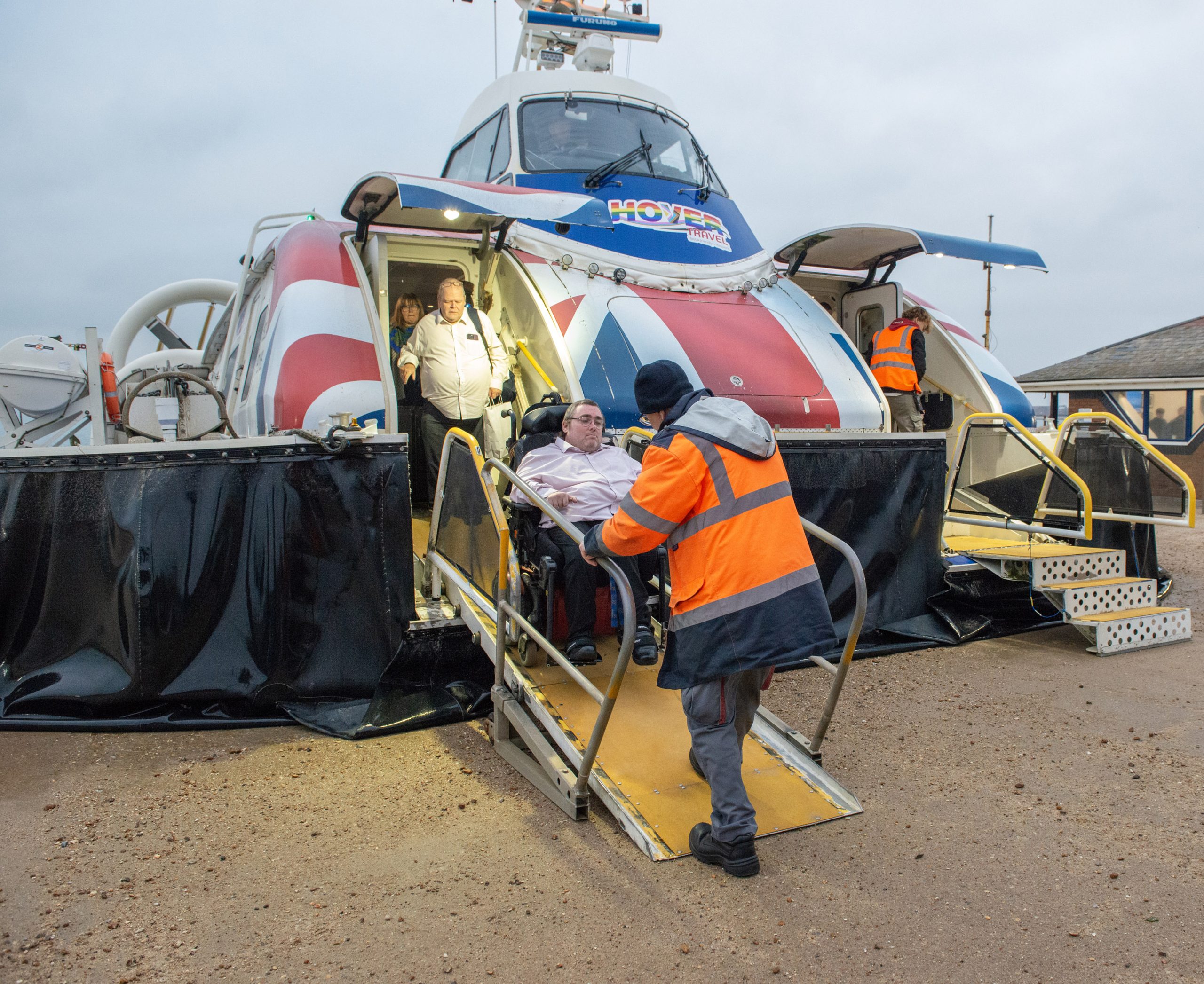 A man in a wheelchair is assisted down a ramp from a Hovertravel hovercraft by a Hovertravel employee at Southsea in Hampshire. Two other people are also leaving the hovercraft.